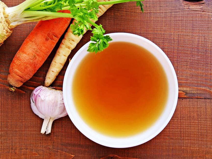Clear bone broth in a white bowl and vegetables on wooden table top view.