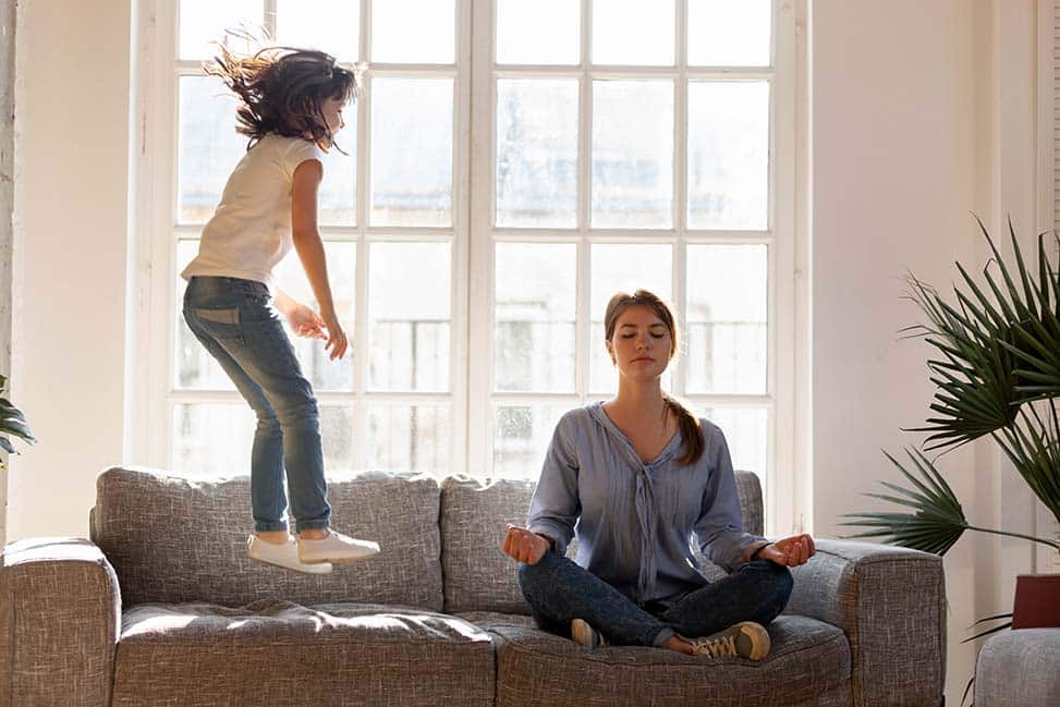Young mom sit on couch at home meditating in lotus position, small daughter have fun jumping near, concentrated calm mother practice yoga relax on sofa ignore playful kid. Stress free concept.