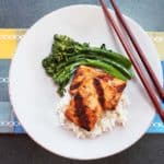 A white plate and burgundy chopsticks against a blue and golden table cloth topped with broccolini, and grilled blackened tofu on a bed of rice.