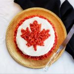 A red watermelon cake with white icing and a Canadian maple leaf using diced strawberries as the main design on a golden plate with a knife.