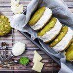 Matcha and white chocolate coconut ice cream sandwiches displayed in a bread pan with parchment paper on a cooling rack placed on a wooden countertop. Around the bread pan is matcha white chocolate cookies, chunks of white chocolate and measuring spoons with matcha and coconut flakes inside two of them. The other two are empty.