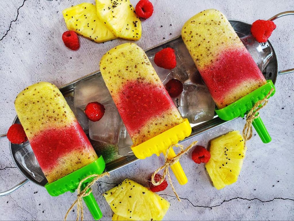 Raspberry and pineapple popsicles in plastic holders with twine tied around the handles. The popsicles are in a metal tray on ice, raspberries and pineapples. The metal tray sits on top of a concrete counter top.