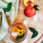 A top-down shot of Peach and Mint Iced Tea in a mason jar garnished with a mint leaf, a flower and a peach. The background is a wooden platter with ice cubes, mint leaves, peach halves and a napkin decorating it.
