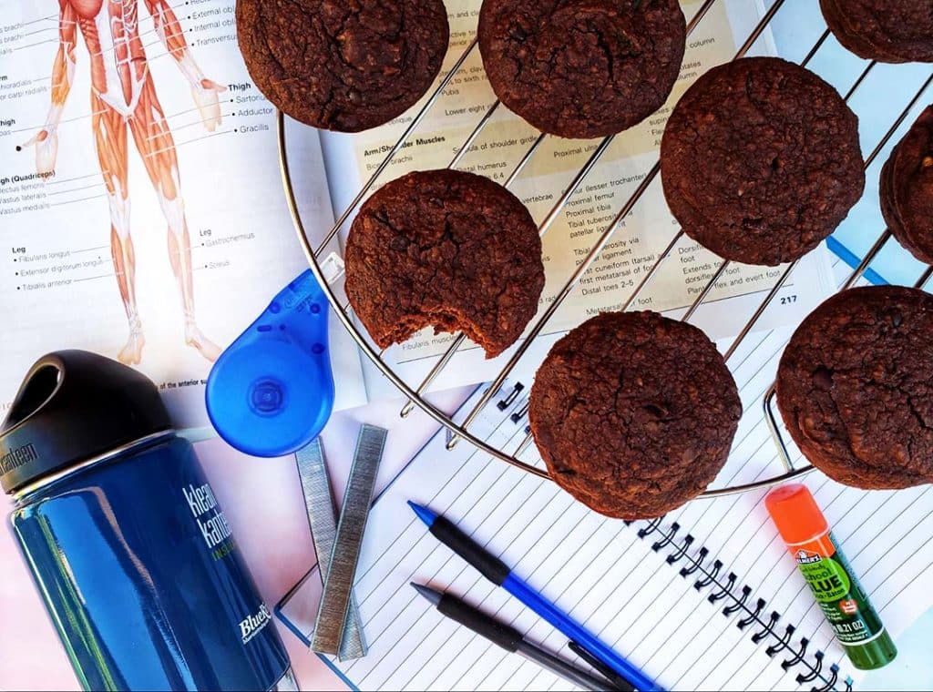 A top-down shot of Chocolate Zucchini Protein Muffins on a circular cooling rack on top of a table with stationery and school supplies on it.