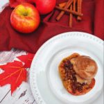 An image of a baked apple crumble served with ice cream on top in a white plate. It is served on a bleached wooden table with red autumn leaves, a red table cloth, two apples and cinnamon sticks as decorations.