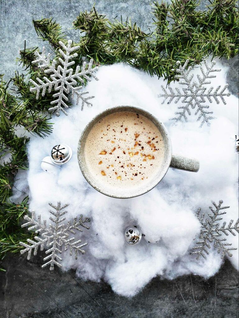 An overhead shot of a latte drink with fake snow, ornaments and evergreen branches as a decoration on a grey concrete-style countertop.
