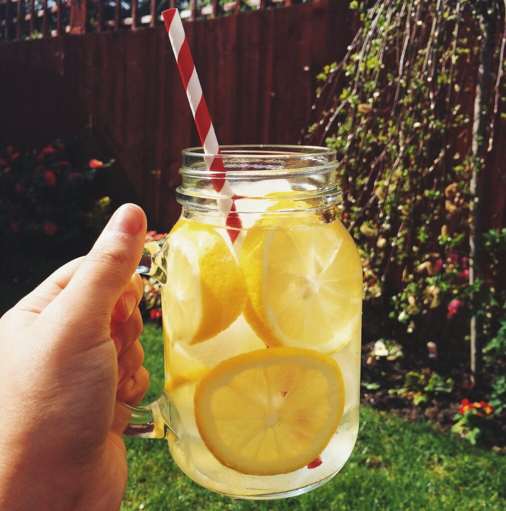 Lemon water held by a hand in a mason jar mug outside in the backyard garden. In the mug, there is a red and white pinstripe paper straw.