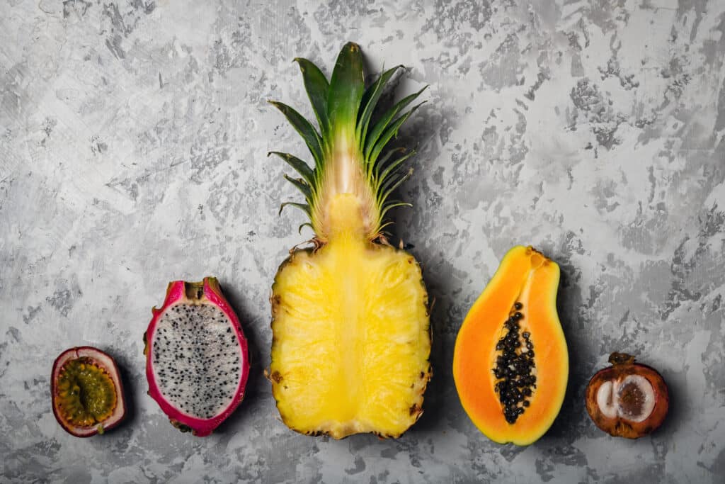 A top-down image of passion fruit, dragon fruit, pineapple, papaya and a mangosteen cut into halves and lined up on a grey concrete countertop.