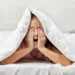 A child laying on their stomach in bed while covering their eyes with their hands with a white blanket over their head.