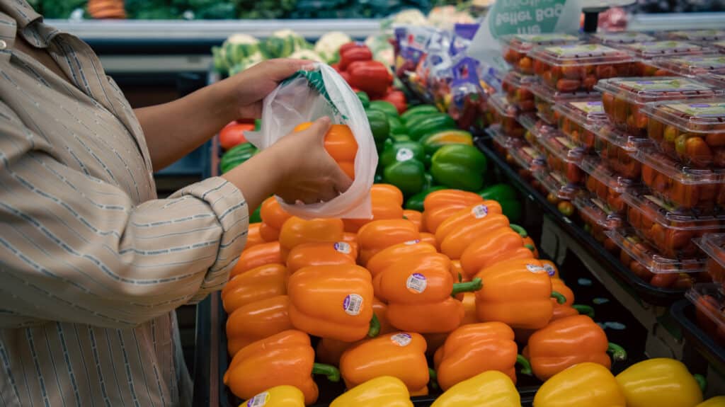 A close-up shot of a customer's hand picking up an orange bell pepper and putting it into a produce bag while shopping at a supermarket.